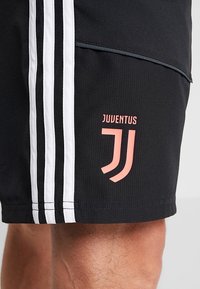 Shorts sportivi neri con strisce verticali bianche e un logo Juventus rosa sulla coscia, indossati da una persona con i peli delle gambe visibili.
