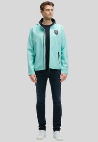 Giacca verde menta con colletto alto, due tasche laterali con zip e una patch con logo sul petto. Indossata sopra una maglietta nera e jeans scuri.