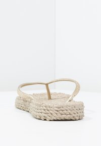 Beige flip-flop med en glittrende rem og en vævet, tekstureret sål. Designet har en flad, bred bund for komfort.