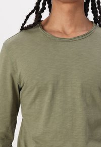 Olivengrünes Langarmshirt aus weichem Stoff; mit rundem Halsausschnitt und sauberen Kanten sowie subtiler Textur im gesamten Shirt.