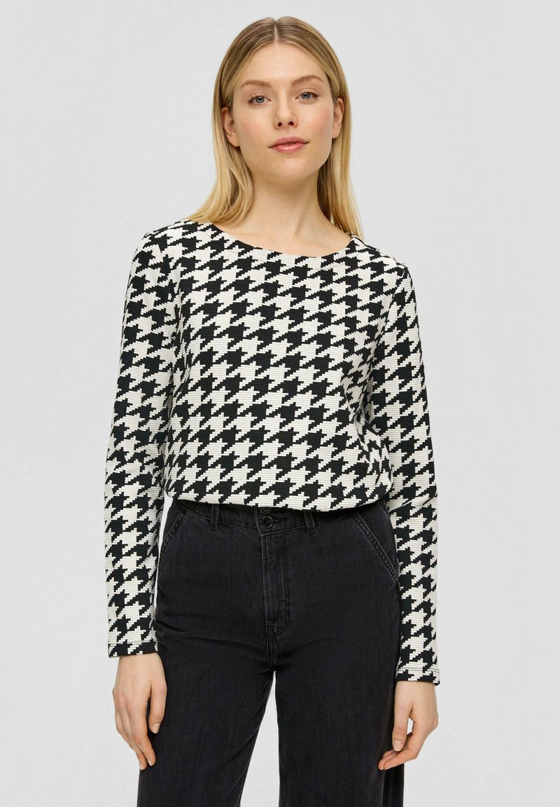 Schwarz-weißes Hahnentristop mit langen Ärmeln und rundem Ausschnitt, kombiniert mit hoch geschnittenen schwarzen Jeans. Glatte Stoffoberfläche.