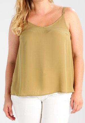 Femme portant un haut ample sans manches de couleur moutarde avec de fines bretelles et un pantalon blanc, devant un fond uni.