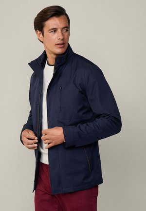 Marineblauwe jacket met een hoge kraag, twee met rits gesloten zakken en bruine accentstiksels. Draag het over een lichte trui en bordeauxrode broek.
