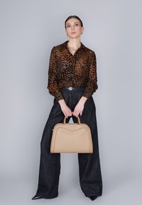 Donna con blusa a stampa leopardata, pantaloni neri a gamba larga e scarpe nere a punta, che tiene una borsa beige contro uno sfondo grigio.