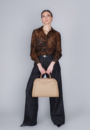 Vrouw in een blouse met luipaardprint, zwarte wijde broek en zwarte puntige schoenen, die een beige handtas vasthoudt tegen een grijze achtergrond.