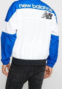 Blå och vit vindjacka med elastisk midja, med "new balance"-logotyp och svarta detaljer på ryggen.