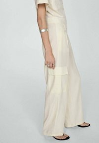 Jumpsuit oversized di colore beige chiaro realizzato in un tessuto morbido e liscio. Presenta un colletto, dettagli sulle tasche ed è abbinato a sandali neri.