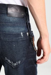 Tmavě modré džíny z denimu se zašlými detaily, koženým štítkem na pásu a kovovým řetězem doplněným hnědým koženým akcentem.