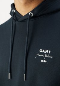 Gros plan d'un sweat à capuche sombre avec des cordons blancs et un texte brodé "GANT American Sportswear 1949" sur la zone de la poitrine.