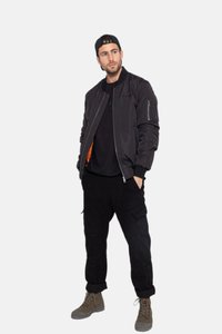 Schwarze Bomberjacke mit Reißverschlusstaschen, über einem dunklen Hemd getragen, kombiniert mit schwarzen Cargo-Hosen und braunen Stiefeln. Das Model steht lässig.