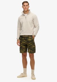 Sudadera cremosa con cremallera y mangas largas, combinada con pantalones cortos cargo camuflados en tonos verdes y oscuros, y zapatillas beige con calcetines a juego.