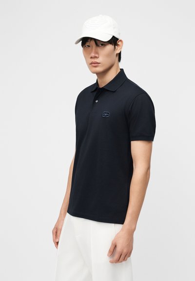 Νέος άνδρας με μαύρο κοντομάνικο πόλο και λευκό παντελόνι, φορώντας λευκό καπέλο, στέκεται μπροστά σε απλό φωτεινό φόντο.