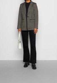 Blazer a quadri bianco e nero con spalle imbottite, abbinato a pantaloni neri svasati e scarpe chunky nere. Tiene una piccola borsa bianca.