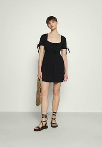 Mulher vestindo um vestido preto curto com mangas atadas, segurando uma bolsa de palha e usando sandálias pretas com atacadores, em pé contra um fundo simples.