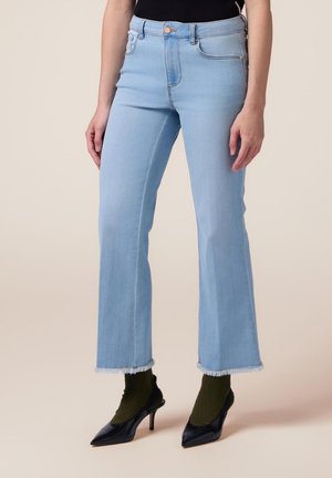 Person iført lyseblå cropped flare jeans med frynser ved kanterne, sorte spidse høje hæle og mørkegrønne sokker på en ensfarvet baggrund.