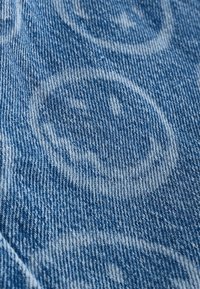 Denim-kanga, joka esittelee sinistä pesua haalistuneella hymiön kuvioinnilla; teksturoitu kudos näkyvissä, luoden rennon ja leikkisän ilmeen.