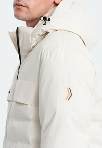 Vit puffjacka med huva, som har en frontdragkedja, en bröstfickslapp och en logotyp-patch med orange och svarta detaljer.