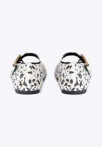 Schwarze und weiße gepunktete flache Schuhe mit durchbrochenem Muster und goldenen Schnallenriemen, von hinten auf einem weißen Hintergrund betrachtet.