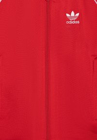 Chaqueta roja con cremallera hecha de tejido texturizado, que presenta el logo blanco de Adidas, dos bolsillos laterales y acentos de canalé a lo largo de los bordes.