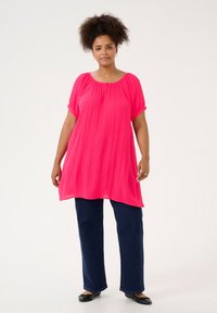 Tunic fucsia brillante con maniche corte arrotolate, scollatura plissée e vestibilità morbida, abbinata a jeans larghi blu scuro e ballerine nere.