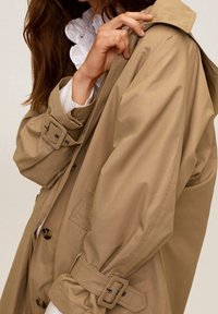 Manteau beige en trench avec un tissu lisse et résistant à l'eau. Il se distingue par un design double boutonnage, de grands boutons et une ceinture avec des accents de boucle.
