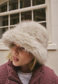 Faux-Fur-Bucket-Hut in hellbeige, mit breiter Krempe und weicher Textur. Getragen mit einer bordeauxfarbenen karierte Jacke.