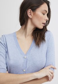Lichtblauwe cardigan met een V-hals, korte mouwen en twee decoratieve knoopaccenten aan de voorkant. Gladde textuur en fijne brei.