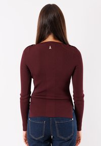 Top lungo a maniche ribbed color bordeaux con un design aderente, caratterizzato da un piccolo logo dorato sul retro e dettagli sulle spalle.