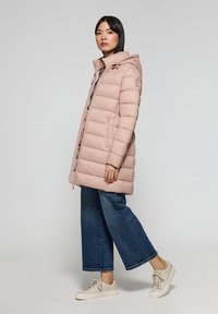 Jasnoróżowy, ocieplany płaszcz z kapturem, z zamkiem błyskawicznym z przodu i kieszeniami bocznymi. W połączeniu z szerokimi niebieskimi dżinsami i beżowymi sneakersami.