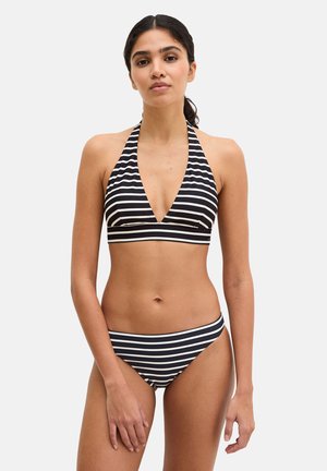 Femme portant un haut de bikini style licou à rayures horizontales noires et blanches et un bas de bikini assorti, debout devant un fond uni.