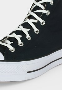 Κοντινή λήψη μαύρου καμβά sneaker υψηλού πάνω μέρους με λευκά κορδόνια, μεταλλικά περβάνια και παχιά λευκή καουτσούκ σόλα με υφασμένη προστασία δακτύλων.