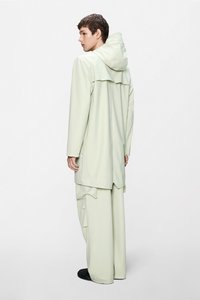 Ανοιχτό πράσινο αδιάβροχο μπουφάν με κουκούλα, μακριά μανίκια και άνετη εφαρμογή, σετ με αντίστοιχο χαλαρό παντελόνι με διακριτικές πτυχώσεις.