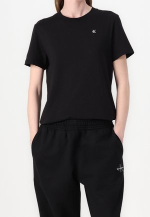 Osoba ubrana w czarną koszulkę z krótkim rękawem z małym logo CK oraz czarne elastyczne spodnie z logo Calvin Klein, ręce w kieszeniach.