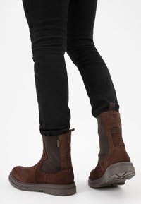 Stivali alla caviglia in suede marrone con pannelli elastici laterali, upper testurizzato e suola di gomma robusta. Gli stivali sono abbinati a pantaloni neri aderenti.