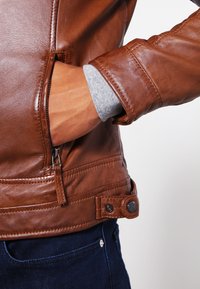 Chaqueta de cuero marrón con un acabado texturizado, bolsillo con cremallera lateral y detalle de botón en la cintura. Tela gris asoma desde el puño.