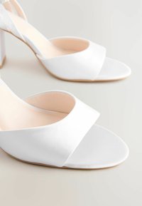 I sandali alti in satin bianco presentano un ampio design a fascetta incrociata, punta aperta e una texture liscia con una soletta nude e un tacco in legno chiaro.
