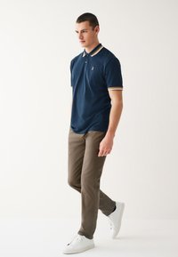 Navy polo shirt with a three-button placket, beige-striped collar and cuffs, paired with light brown pants and white sneakers.