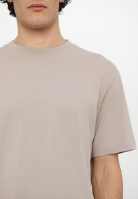 Beżowa koszulka z okrągłym dekoltem, wykonana z miękkiej bawełny. Posiada krótkie rękawy i luźny krój, z minimalnymi szwami i bez wzorów.