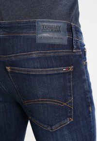 Jeans de mezclilla azul oscuro con costuras naranjas, que cuentan con un parche de cuero etiquetado "TOMMY JEANS". Incluyen bolsillos traseros con costuras en curva.