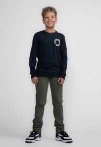 Marineblauwe longsleeve shirt met witte logodetails, gecombineerd met olijfgroene broek en zwart-witte sneakers. Zachte katoenen stof.