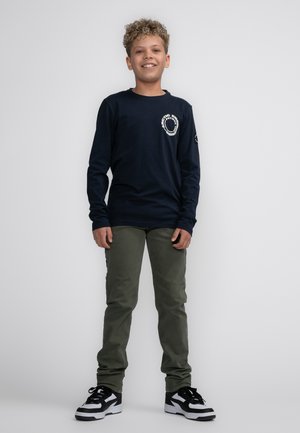 Marineblauwe longsleeve shirt met witte logodetails, gecombineerd met olijfgroene broek en zwart-witte sneakers. Zachte katoenen stof.