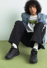Schwarze Sportschuhe mit runden Sohlen, ausgestattet mit Schnürsenkeln und blauen Akzenten, kombiniert mit schwarzen Hosen und einer Jeansjacke. Sitzt auf grünem Boden.