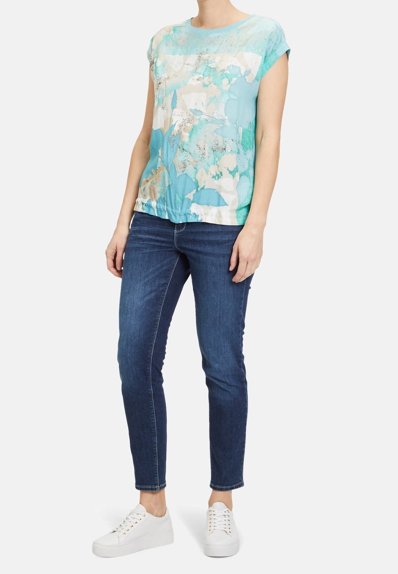 Mujer con una blusa corta de manga azul con estampado floral, jeans ajustados de color azul oscuro y zapatillas blancas con cordones, de pie frente a un fondo blanco.
