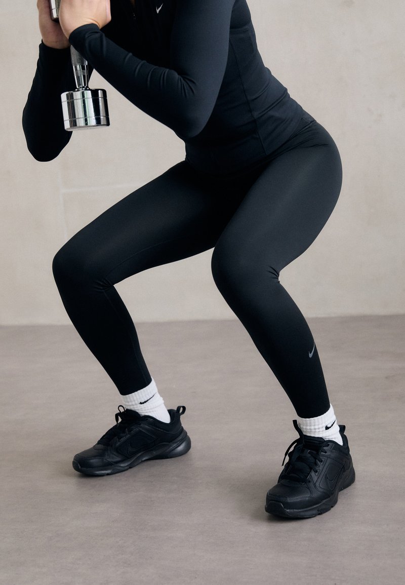 Leggings sportivi neri e top a maniche lunghe con un design aderente, abbinati a scarpe Nike nere e calzini bianchi, mentre tiene un manubrio d'argento.