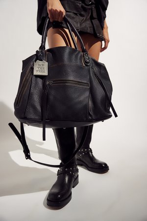 Personne tenant un grand sac en cuir noir avec des poches avant à fermeture éclair et une plaque métallique portant l'inscription « WE THE FREE », portant des bottes en cuir noir cloutées.
