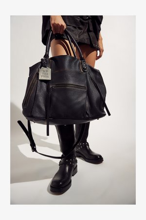 Persona che tiene una grande borsa nera in pelle con tasche frontali con zip e una targhetta metallica con la scritta "WE THE FREE", indossando stivali neri in pelle con borchie.