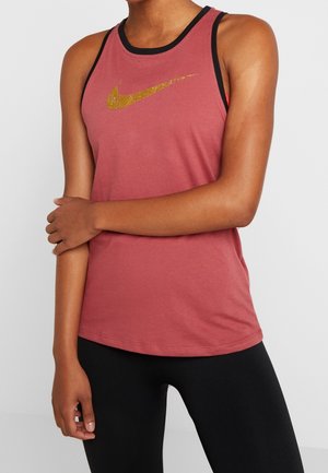 Personne portant un débardeur rose foncé sans manches avec un logo Nike doré et des leggings noirs, debout avec un bras croisé sur l'autre.
