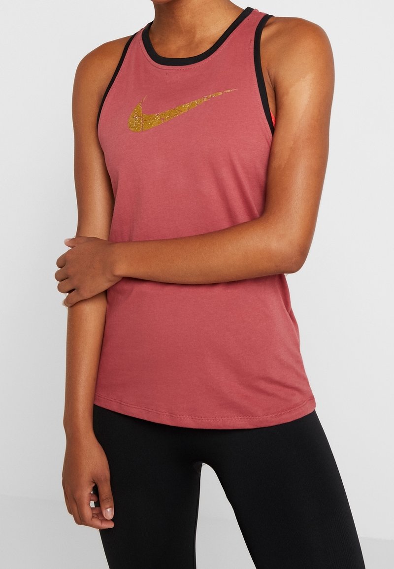 Personne portant un débardeur rose foncé sans manches avec un logo Nike doré et des leggings noirs, debout avec un bras croisé sur l'autre.