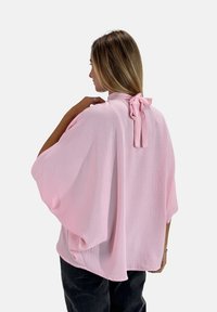 Blusa rosa chiaro con una vestibilità morbida, spalle cadenti e dettaglio a fiocco sul retro. Il tessuto ha una texture stropicciata.