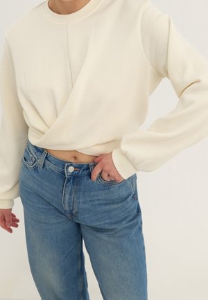 Sweatshirt crème avec un col rond et un détail de torsion sur le côté, associé à un jean taille haute bleu clair, avec une fermeture à bouton.